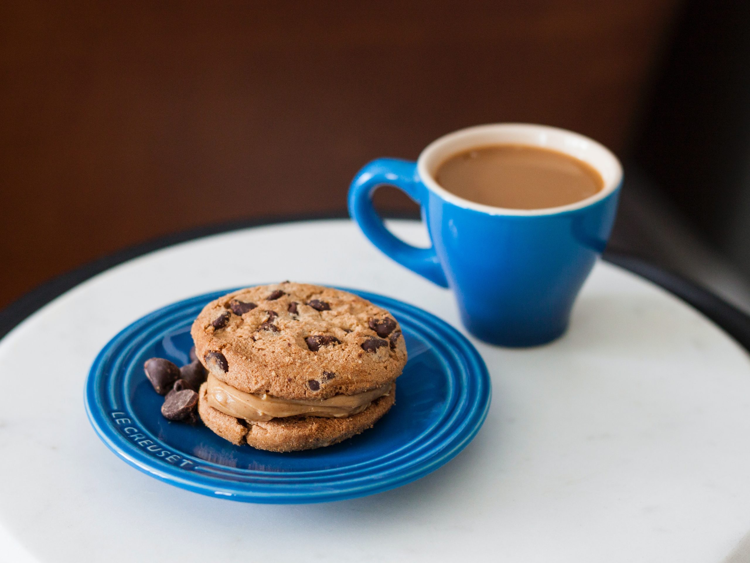 A small blue mug with milky coffee next to a blue plate with chocolate chip cookies.