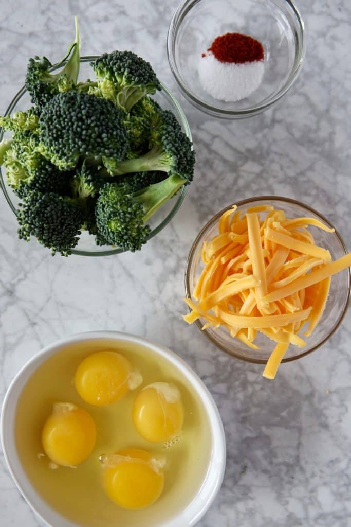 ingredients in bowls displayed on a marble counter, including 4 eggs, shredded cheddar cheese, broccoli florets, and seasonings.
