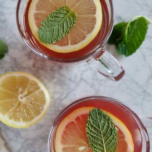 View of two glass mugs with red tea, garnished with lemon slices and mint leaves.