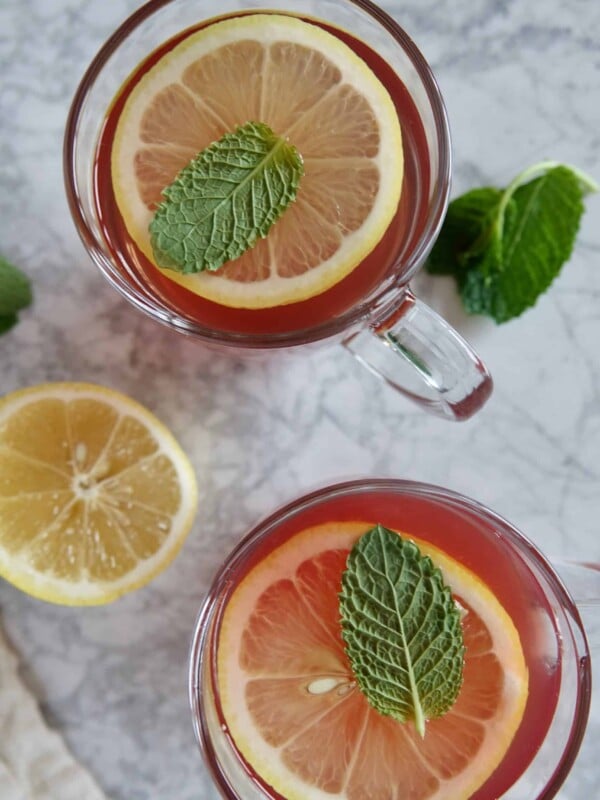View of two glass mugs with red tea, garnished with lemon slices and mint leaves.