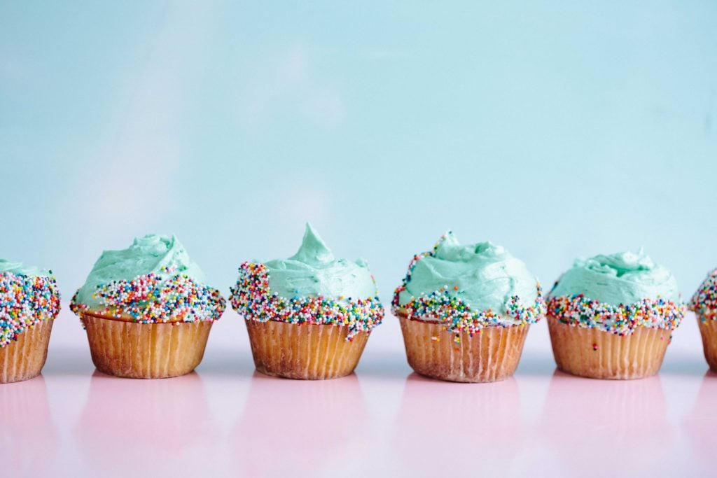 six cupcakes with teal frosting and sprinkles lined up on a pink countertop. 