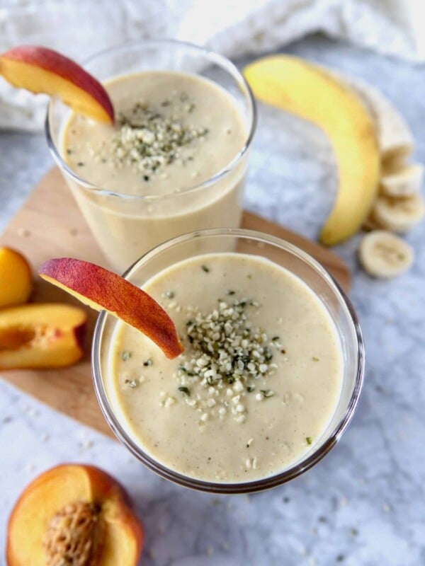 Two glasses filled with smoothies on a marble counter. Peached and bananas are displayed as well.