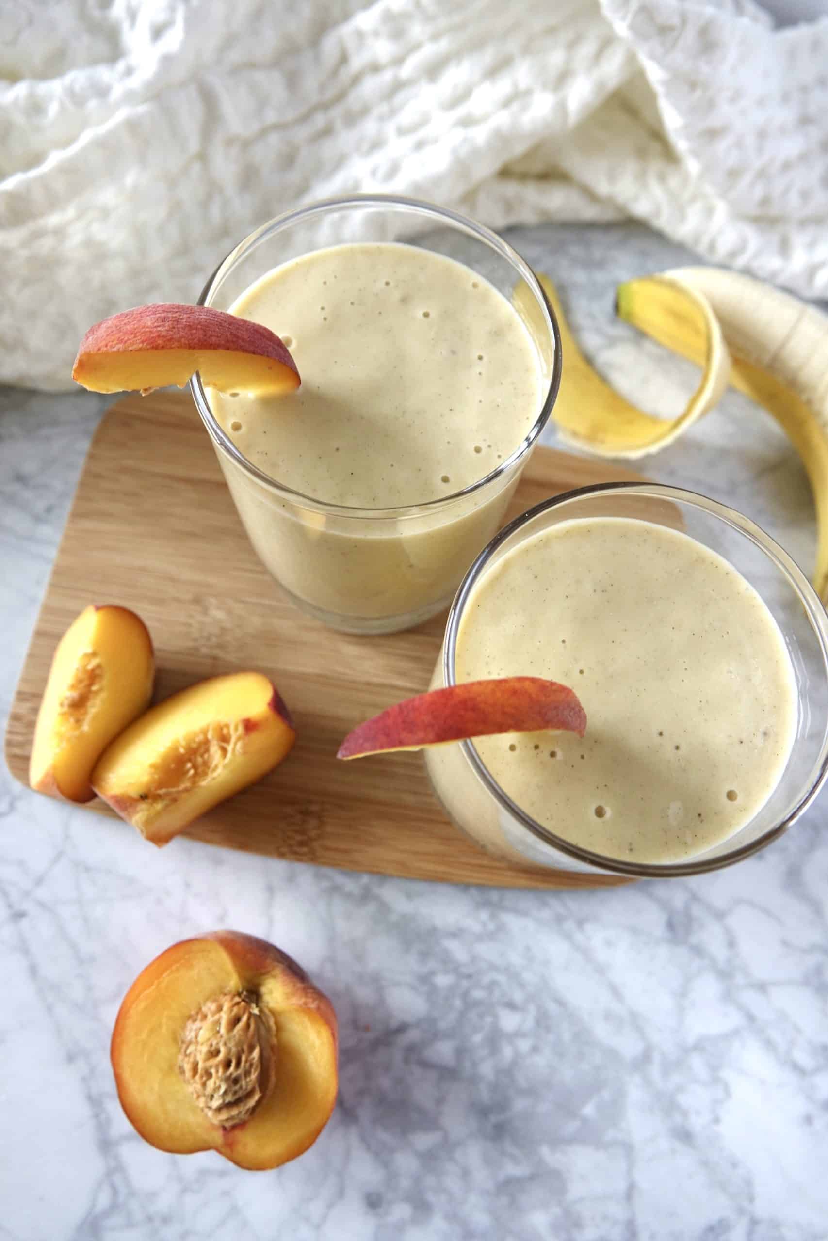 This banana peach smoothie is creamy, refreshing, and made with just 5 ingredients! An easy snack or breakfast ready in 3 minutes. Two glasses with banana peach smoothies, garnished with peach slices. Placed on a white marble counter.