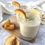 Two glasses with light orange smoothies, garnished with hemp seeds and peach slices. Displayed on marble counter.