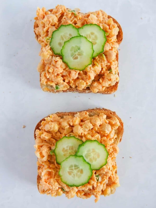 Open faced sandwich with chickpea salad sandwich mixture and sliced cucumbers, sitting directly on white marble countertop