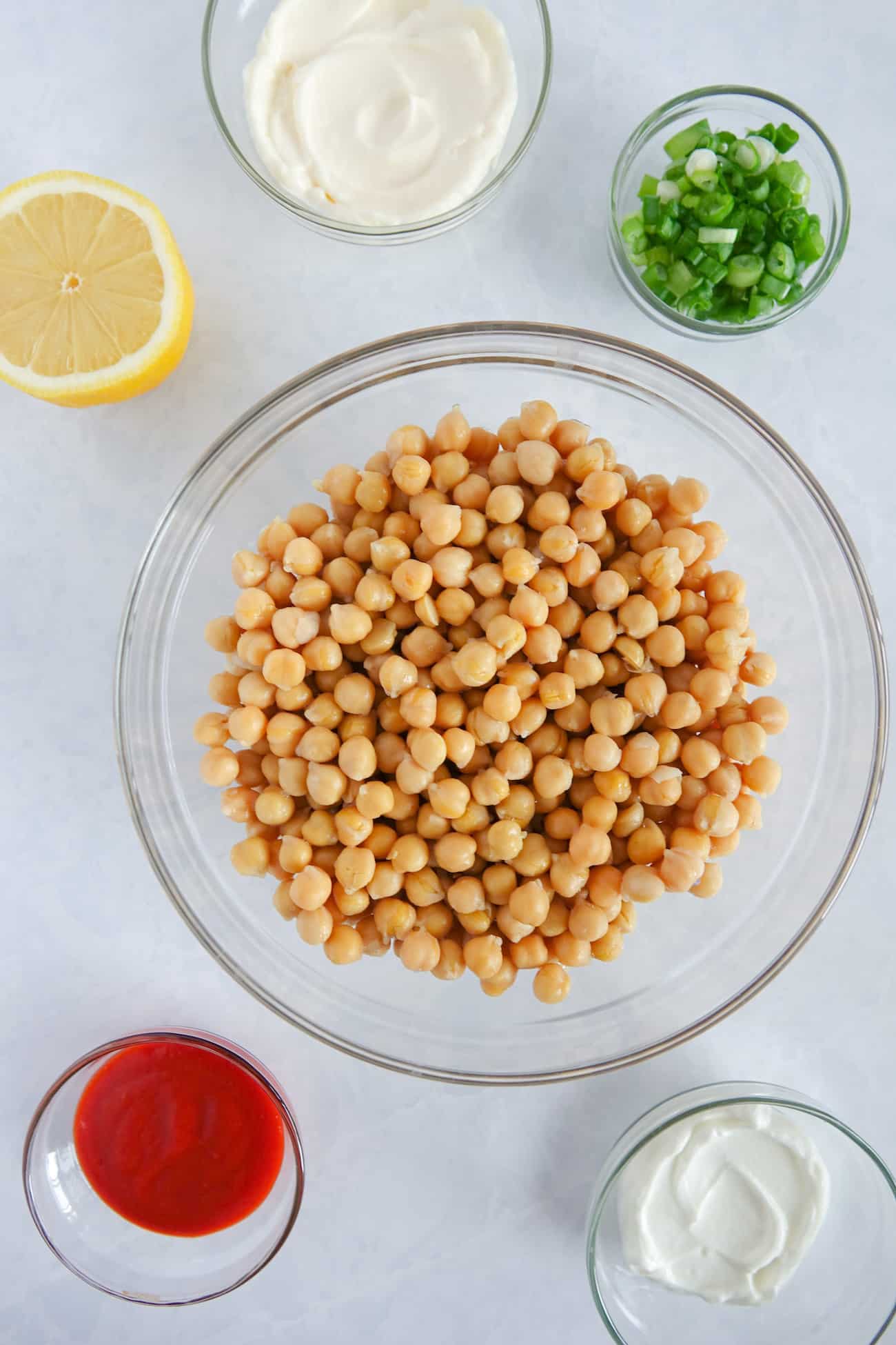 Ingredients for spicy chickpea mash in glass bowls on white marble counter, including canned chickpeas, sriracha, mayonnaise, greek yogurt, half a lemon, and sliced green onion.