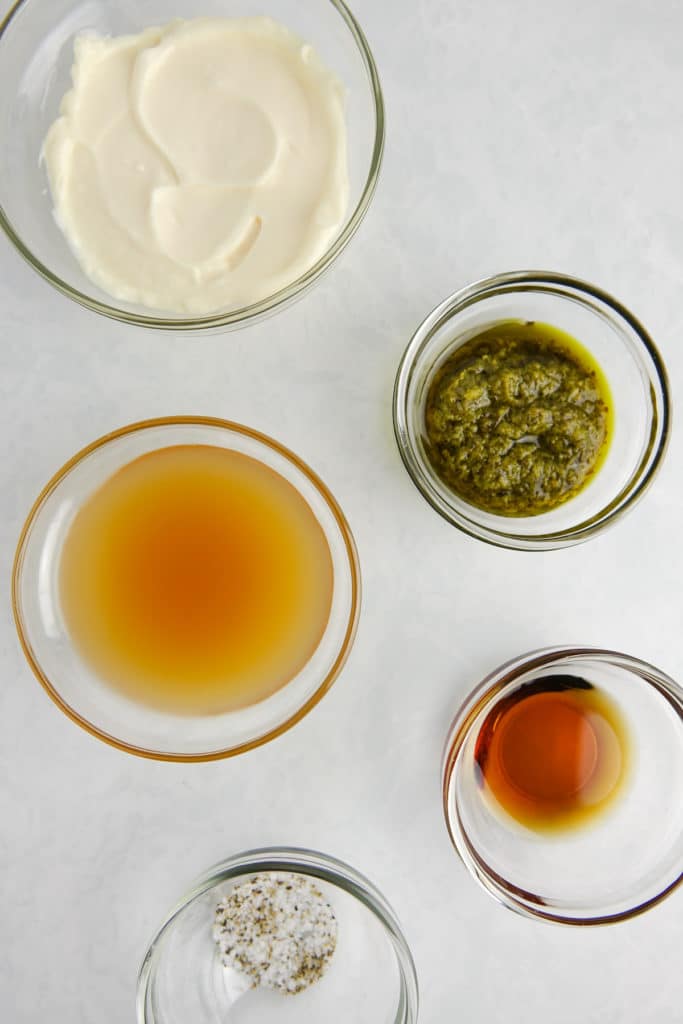 Ingredients in glass bowls on a white marble counter, including mayonnaise, basil pesto, apple cider vinegar, maple syrup, and salt and pepper.