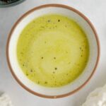 Green salad dressing in a white ceramic bowl on a white marble counter, surrounded by a white kitchen towel and a bowl of broccoli