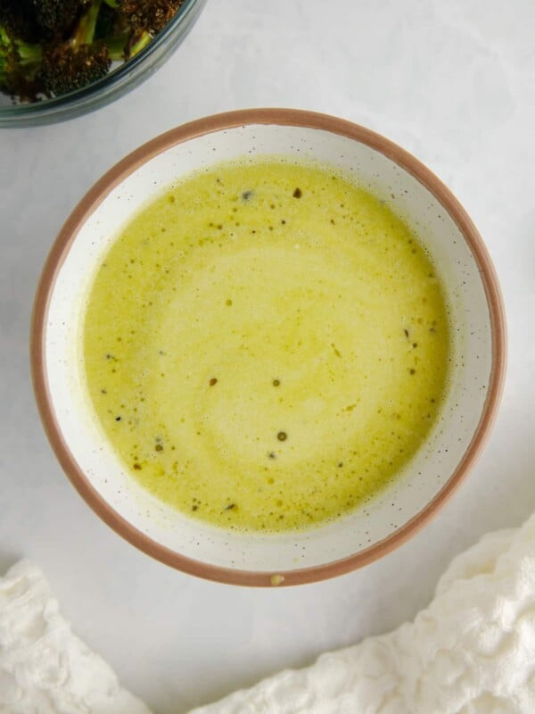 Green salad dressing in a white ceramic bowl on a white marble counter, surrounded by a white kitchen towel and a bowl of broccoli