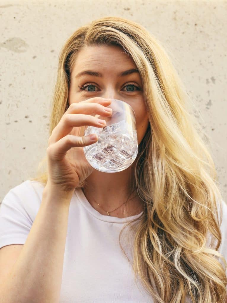 Woman in white shirt drinking a glass of    water outside, in front of a beige wall.