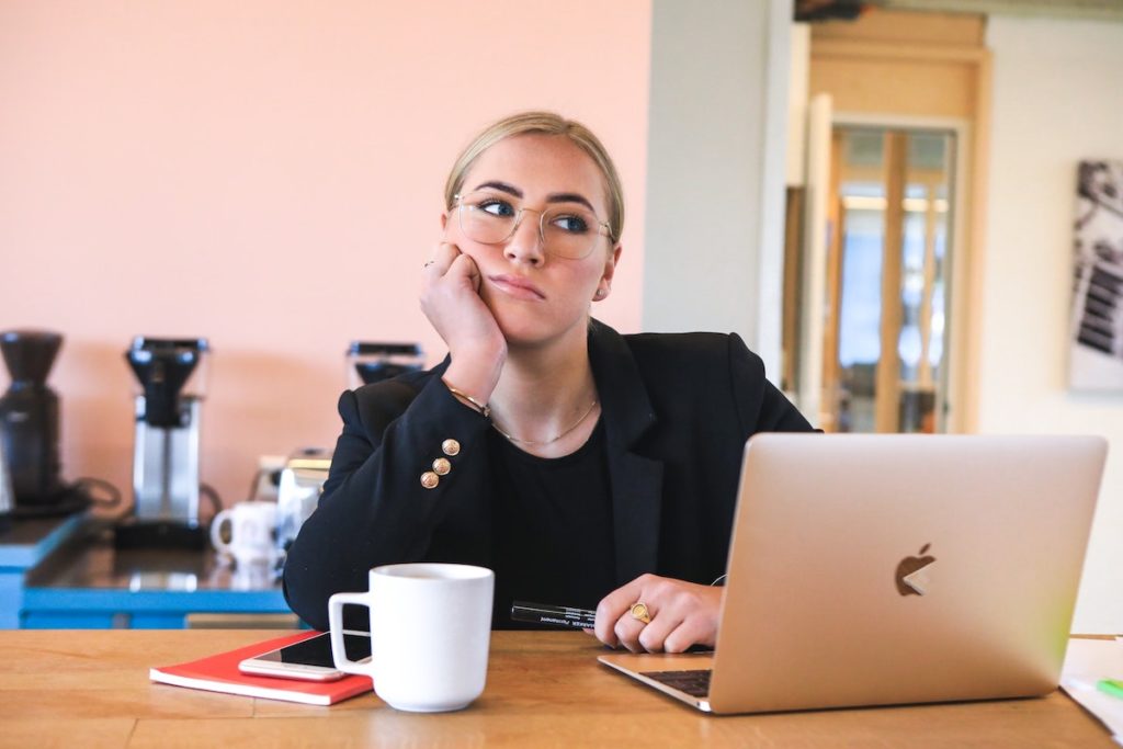 Alt-text: A woman in a professional black suit looking bored in front of her laptop.