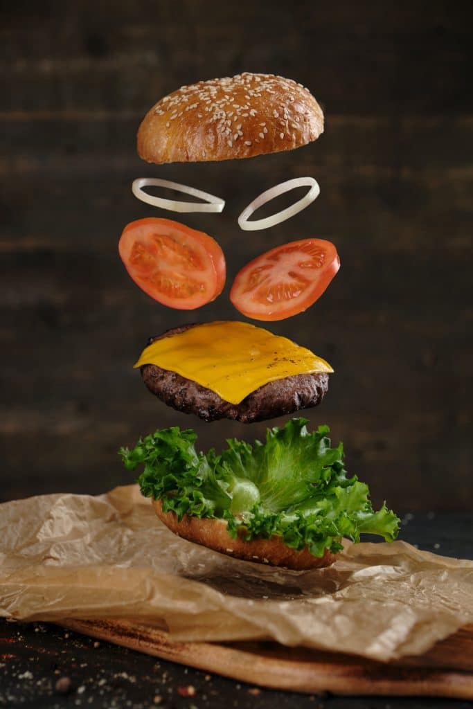 Alt-text: An artistic shot of a cheeseburger with all the toppings flying through the air.