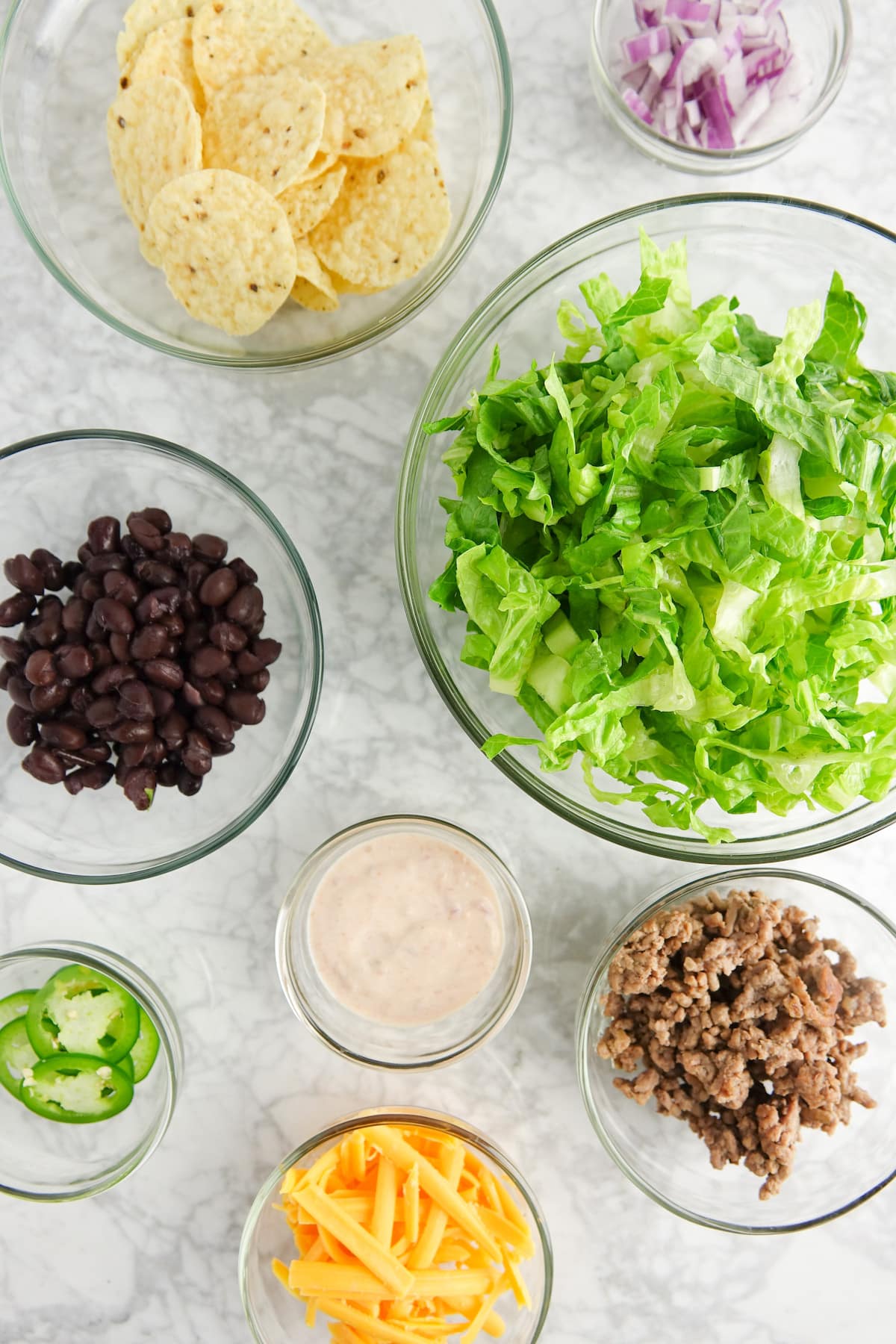 Ingredients displayed in glass bowls on a white marble countertop; tortilla chips, red onion, romaine lettuce, black beans, salad dressing, jalapeno slices, shredded cheese and cooked ground beef.