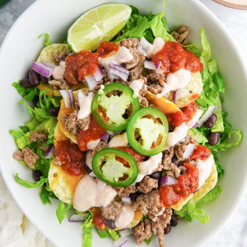 White bowl with salad that includes lettuce, ground beef, black beans, tortilla chips, melted cheese, jalapeno slices, lime wedge, and salad dressing.