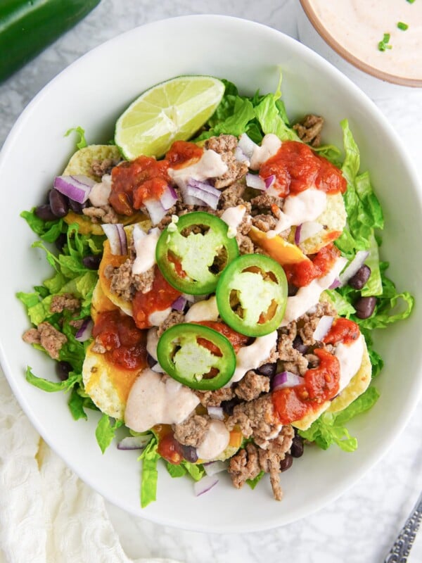 White bowl with salad that includes lettuce, ground beef, black beans, tortilla chips, melted cheese, jalapeno slices, lime wedge, and salad dressing.