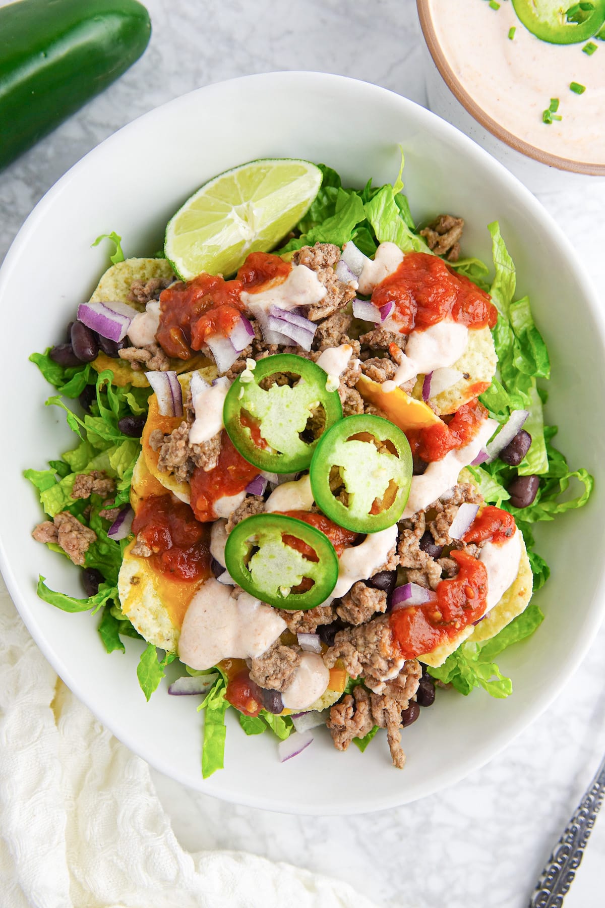 White bowl with salad that includes lettuce, ground beef, black beans, tortilla chips, melted cheese, jalapeno slices, lime wedge, and salad dressing.