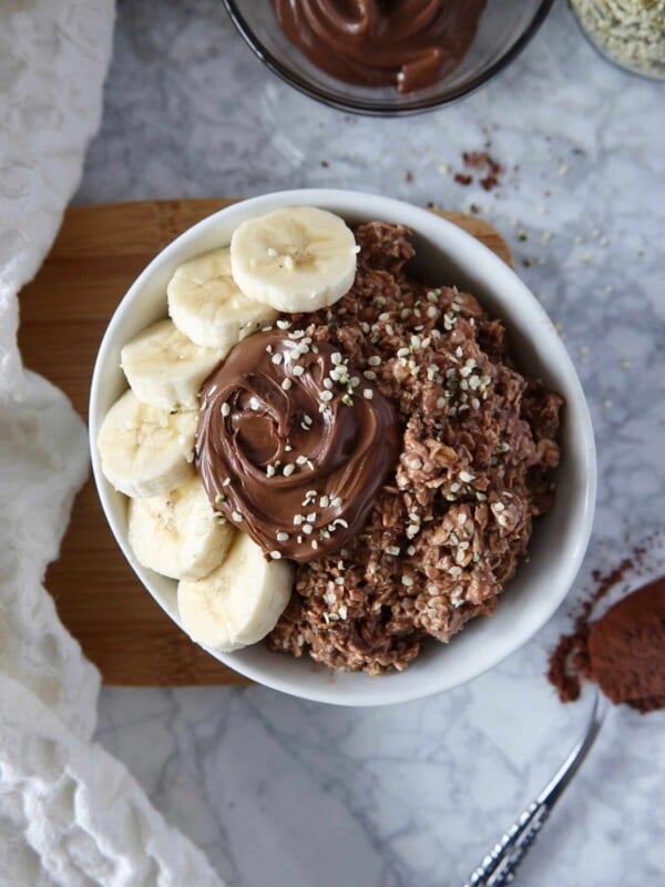 Overhead photos of chocolate overnight oats in a white bowl topped with nutella and banana. Bowl on a white marble counter with ingredients displayed.