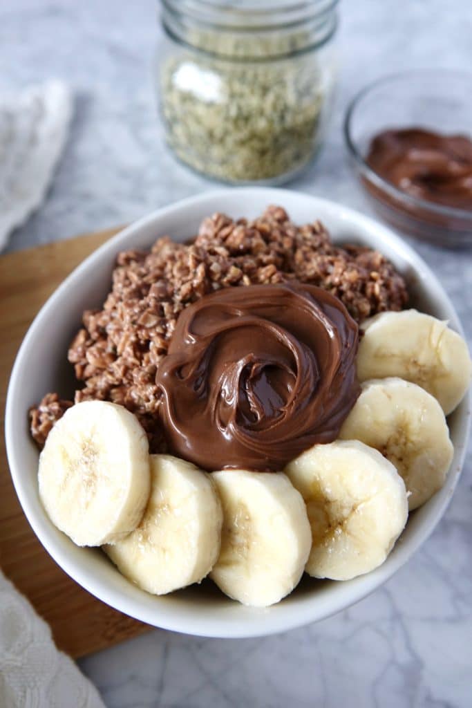 White bowl of chocolate oats topped with nutella and banana. Hemp seeds and nutella displayed on counter behind the bowl of oats.