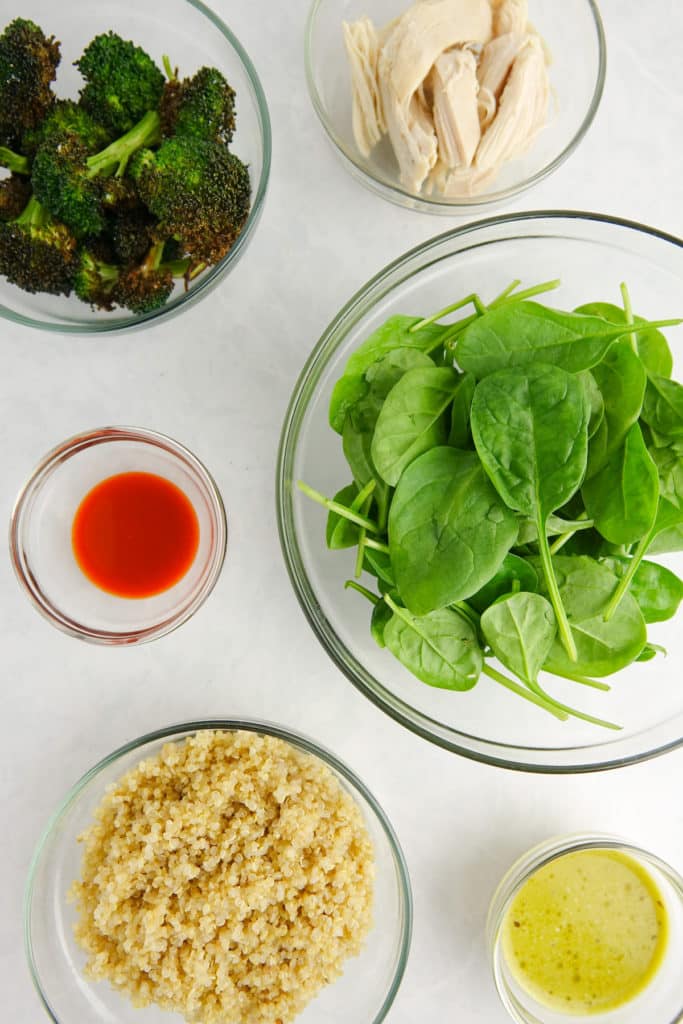 Ingredients displayed in glass bowls on a white marble counter - roasted broccoli, shredded chicken, hot sauce, baby spinach, cooked quinoa and green salad dressing.