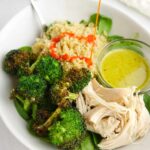 White bowl with cooked quinoa, roasted broccoli, cooked chicken, baby spinach and green salad dressing, drizzled with bright red buffalo sauce.