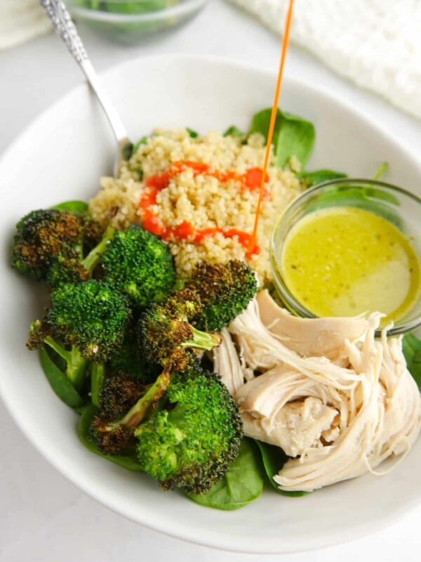 White bowl with cooked quinoa, roasted broccoli, cooked chicken, baby spinach and green salad dressing, drizzled with bright red buffalo sauce.