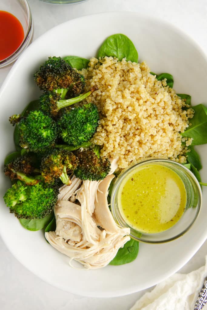 Bowl with spinach, quinoa, roasted broccoli, cooked chicken and a green salad dressing on a white marble countertop