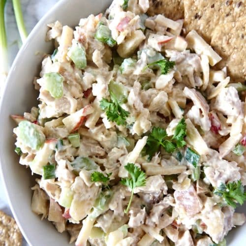 Chicken salad mixture in a white bowl with seed crackers, garnished with lemon, green onion and parsley.