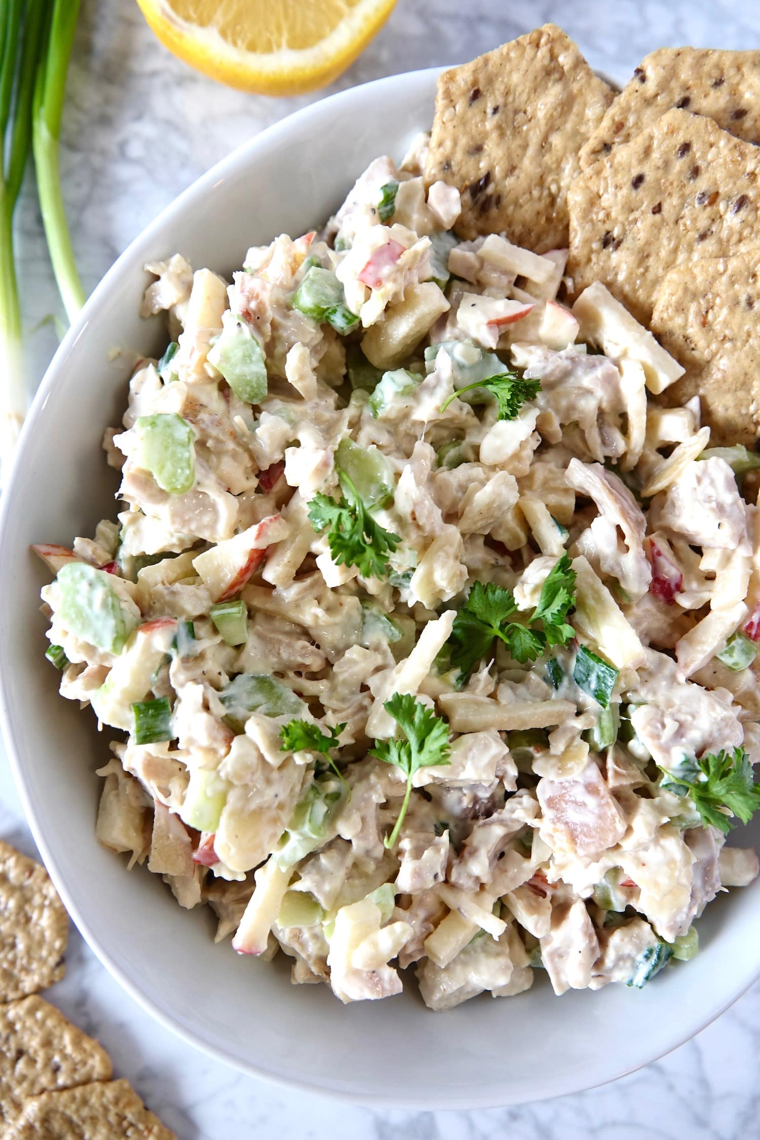 Chicken salad mixture in a white bowl with seed crackers, garnished with lemon, green onion and parsley.