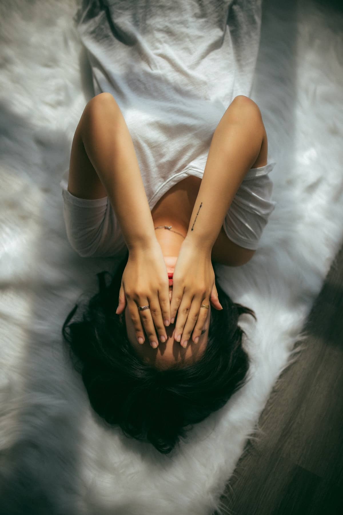 woman laying on bed with white sheets, covering her face with her hands