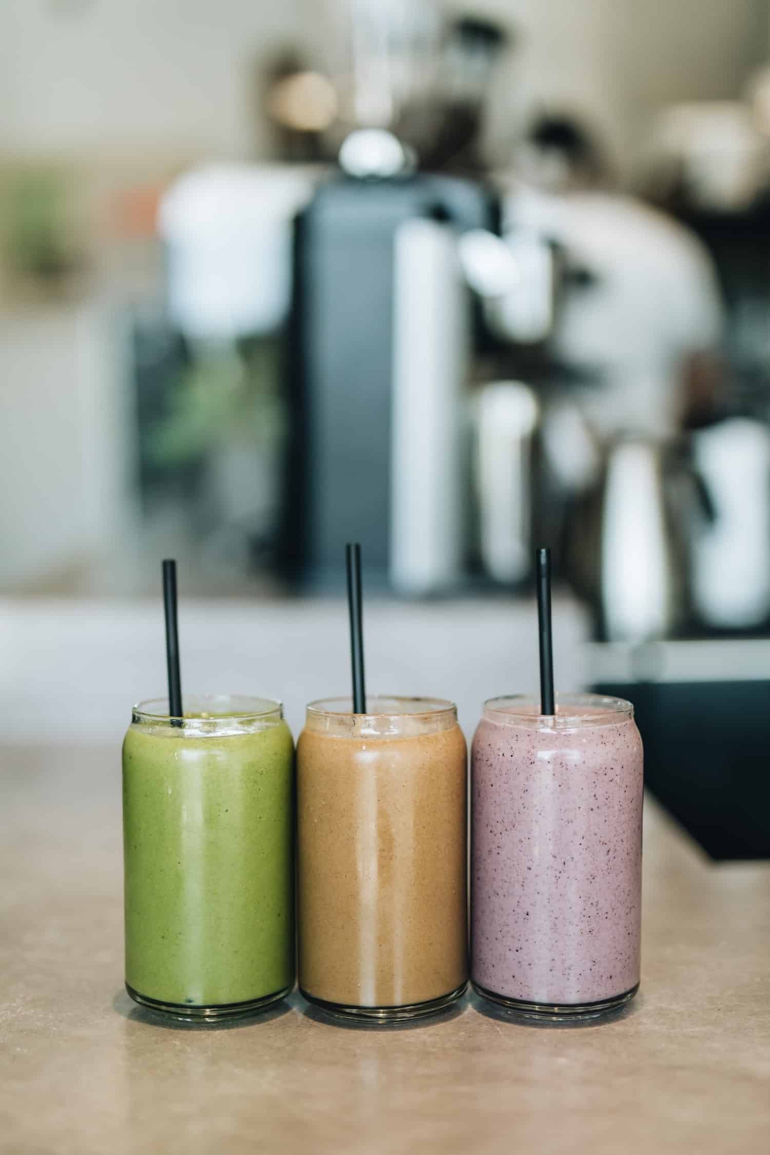 Three smoothies in glass cups with straws, sitting on a counter. Green smoothie, brown smoothie, and purple smoothie are displayed.