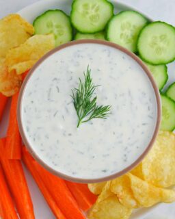 Greek yogurt dill pickle dip presented in a brown bowl on a plate with chopped veggies and potato chips. Food is on a white marble countertop.