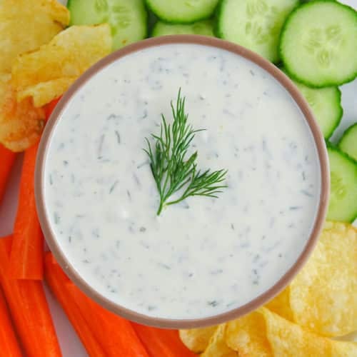 Greek yogurt dill pickle dip presented in a brown bowl on a plate with chopped veggies and potato chips. Food is on a white marble countertop.