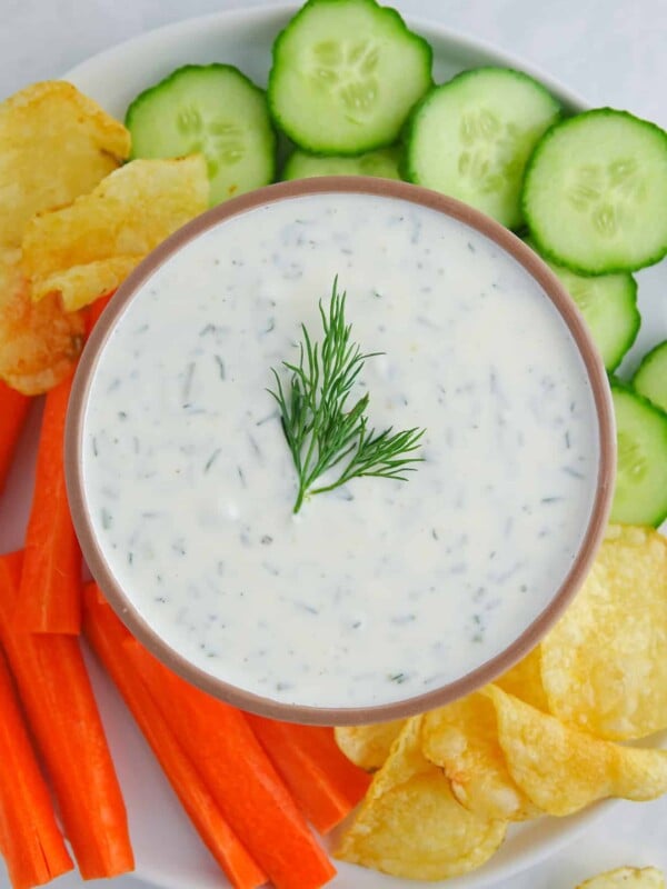Greek yogurt dill pickle dip presented in a brown bowl on a plate with chopped veggies and potato chips. Food is on a white marble countertop.