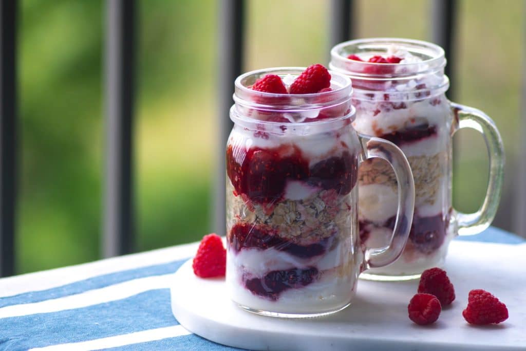two glass jars of layered yogurt and granola with raspberries and raspberry jam.