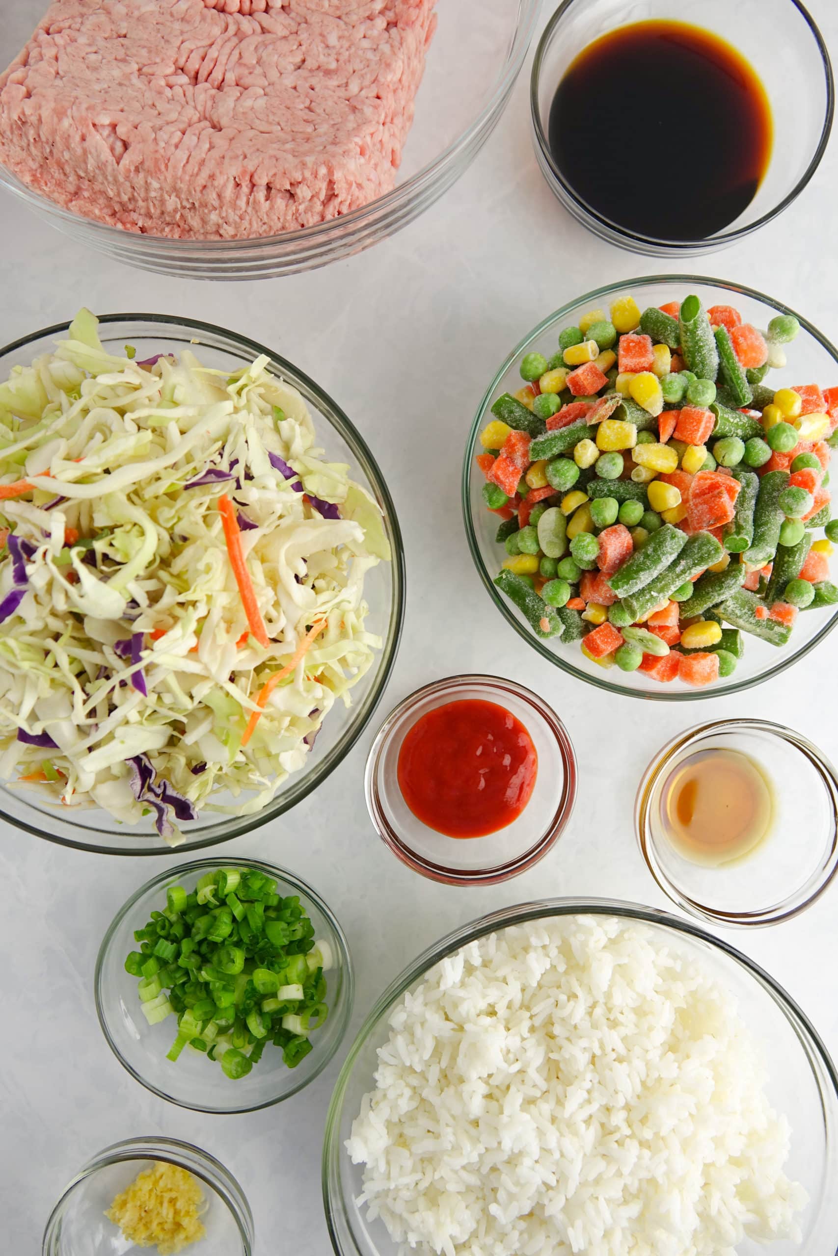 Ingredients for unstuffed egg rolls in a bowl displayed in clear glass bowls on a white marble counter: ground pork, soy sauce, coleslaw mix, frozen vegetables, sriracha, sesame oil, garlic, green onions and cooked rice.