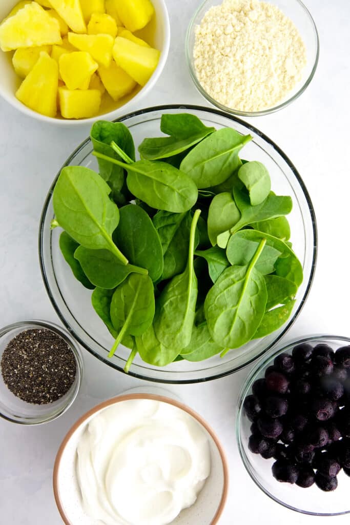 Ingredients for a blueberry pineapple smoothie displayed on a white marble counter in glass bowls. Ingredients include frozen blueberries, frozen pineapple, spinach, yogurt, protein powder, and chia seeds.