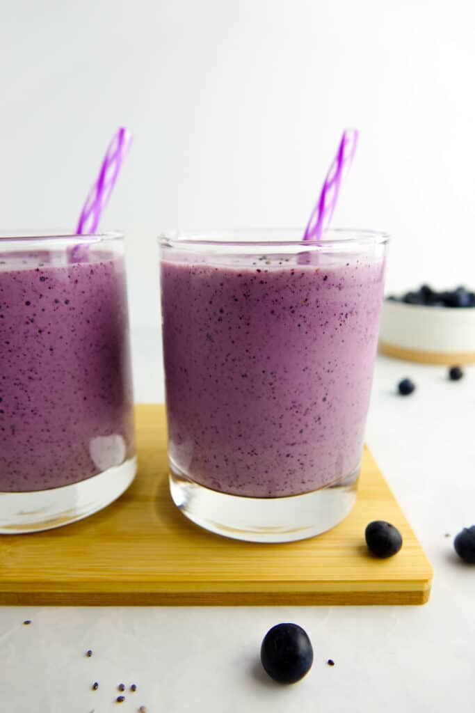 two blueberry smoothies displayed in clear glasses with a straw. Smoothies are placed on a small wood board on a white marble countertop, which also has some chia seeds and blueberries sprinkled around.