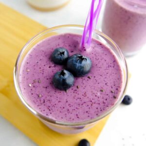 Two glasses containing a purple smoothie and garnished with a striped purple straw and fresh blueberries. Food placed on wood board on white marble countertop.