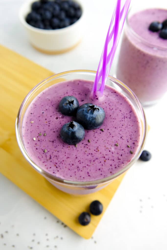 Two glasses containing a purple smoothie and garnished with a striped purple straw and fresh blueberries. Food placed on wood board on white marble countertop.