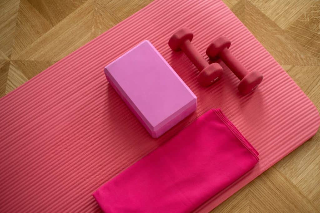 Exercise equipment including dumbbells, a yoga block and a towel placed on a pink yoga mat.