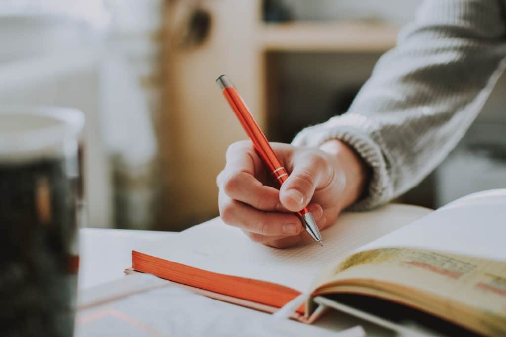 Woman holding an orange pen writing in a notebook.