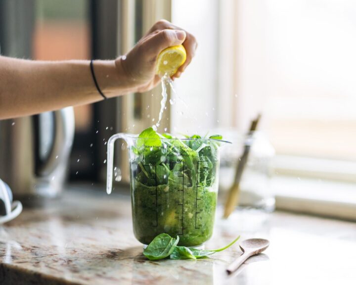 Woman squeezing lemon into a blender containing greens on a marble counter in a kitchen.