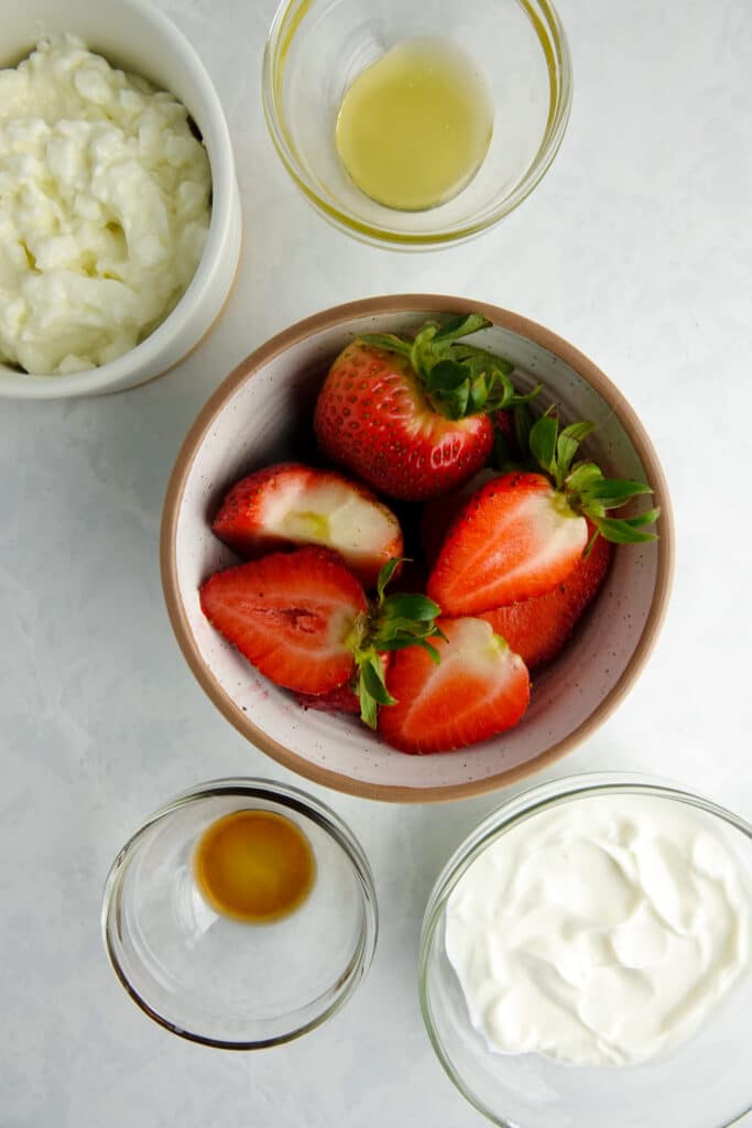 Ingredients for a healthy blended cheesecake bowl in bowls on a white counter - including honey, cottage cheese, greek yogurt, vanilla extract and strawberries.