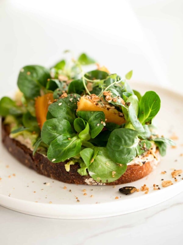 Slice of toast on a white plate topped with ricotta cheese and greens.