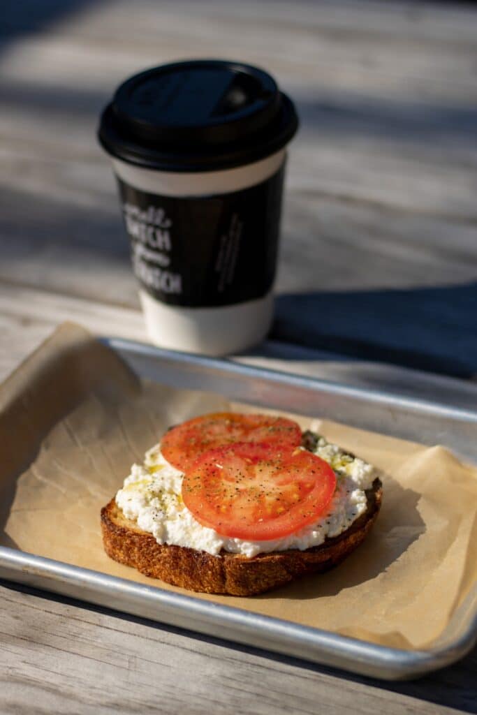 Slice of toast with cottage cheese and two tomato slices on a bench outside with a to-go cup of coffee.