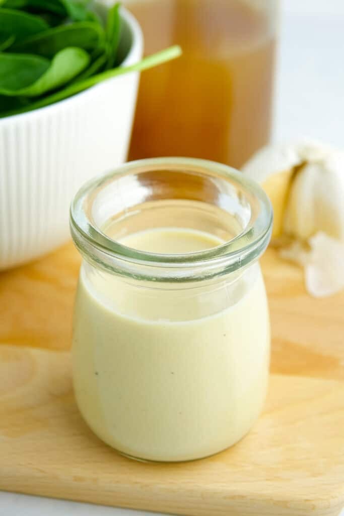 Small glass container filled with hummus salad dressing displayed on a wooden board with greens, garlic and vinegar.