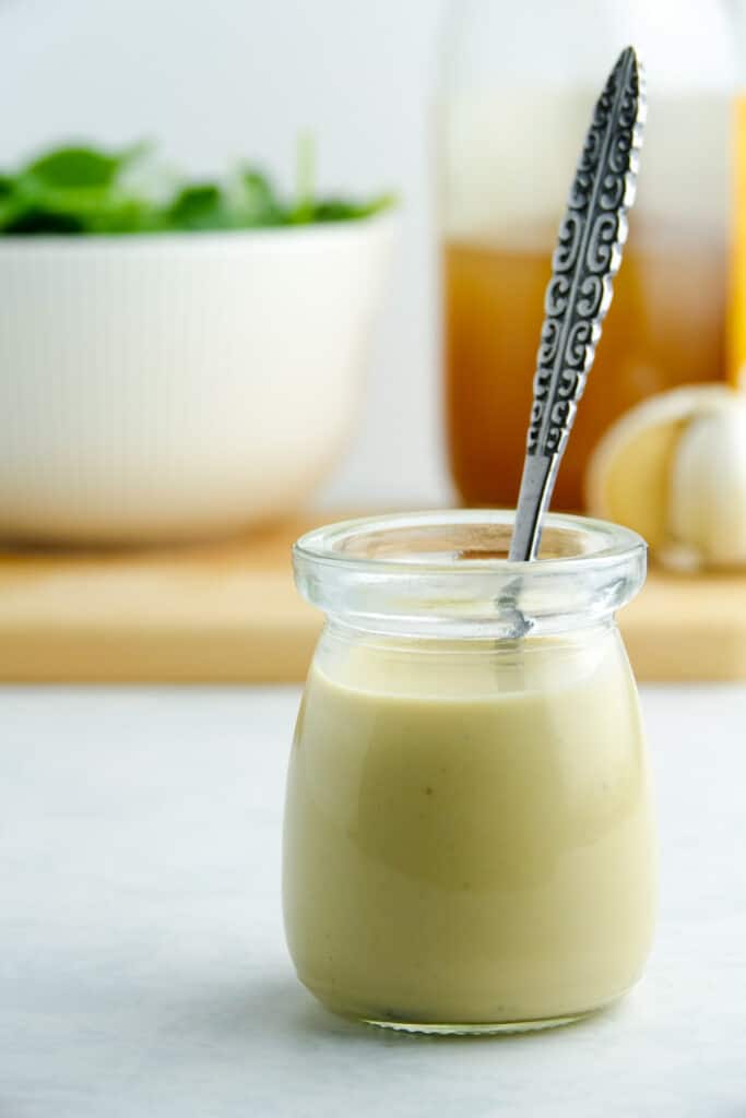 Hummus salad dressing in a small glass container with a metal spoon, displayed on a white counter.