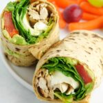 Pesto chicken wrap cut in half and displayed on a white plate with carrots and grapes.
