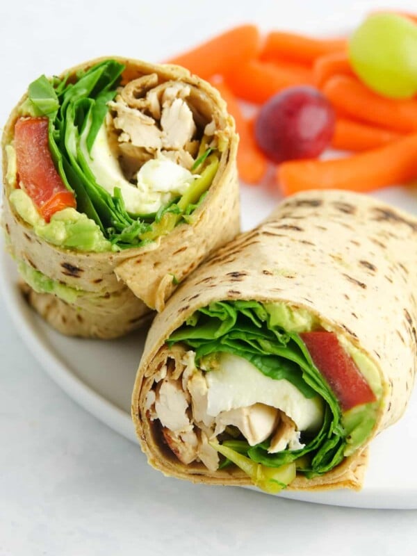 Pesto chicken wrap cut in half and displayed on a white plate with carrots and grapes.