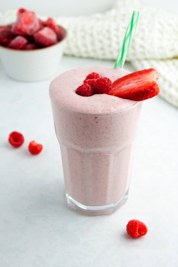 Pink smoothie with strawberries and raspberries in a glass with a green striped straw, displayed on a white marble counter.